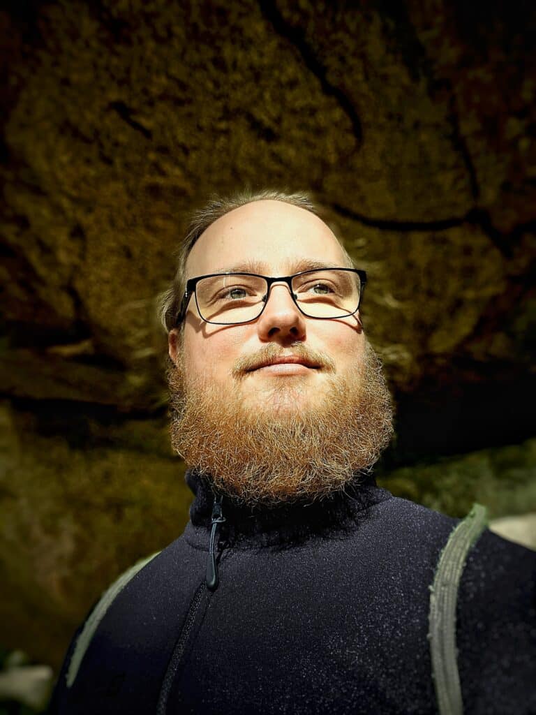 Low-angle portrait of a man with glasses and a full reddish-brown beard illuminated by sunlight, standing beneath a rocky overhang or cave formation.